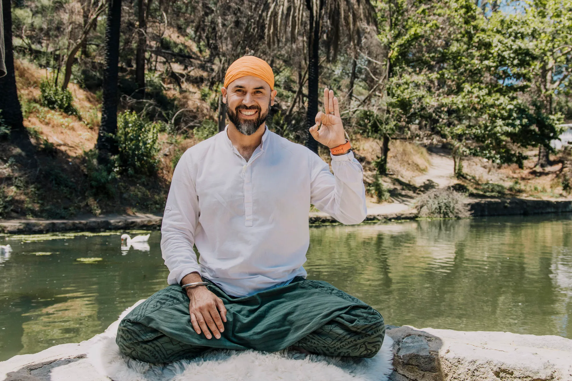 Italo sentado junto al agua, sonriendo, con una mano levantada.